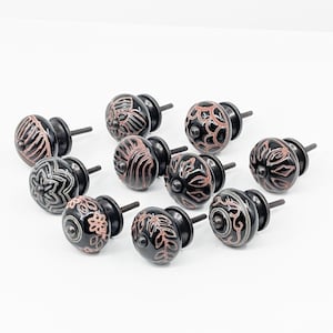 Op de afbeelding: Tien zwarte keramische kastknoppen met ingewikkelde bloemmotieven in koperkleur. De knoppen hebben een zilveren metalen schroefbevestiging.