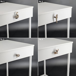 May include: Four white side tables with decorative drawer pulls. Each table has a different antique brass pull: a swan, a parrot, a leaf, and a deer. The tables have white legs and a white tabletop.