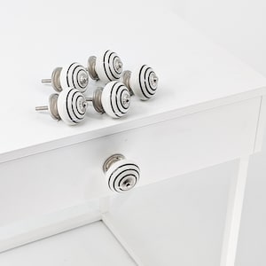 Puede incluir: Conjunto de seis pomos de cajón de cerámica blanca con rayas negras. Los pomos tienen un acabado plateado y se muestran sobre una superficie blanca.