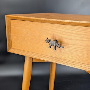 May include: A light-colored wooden nightstand with a drawer featuring a decorative dinosaur-shaped handle. The nightstand has tapered legs and a simple, modern design. The dinosaur handle is dark gray.