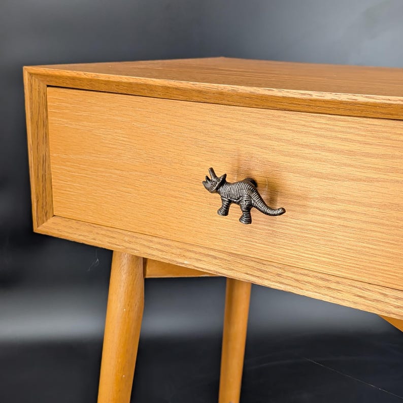 May include: A light-colored wooden nightstand with a drawer featuring a decorative dinosaur-shaped handle. The nightstand has tapered legs and a simple, modern design. The dinosaur handle is dark gray.
