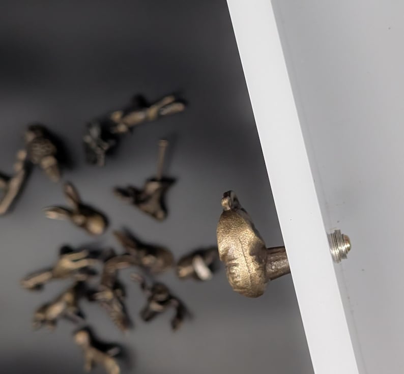 May include: Close-up of a bronze-colored cabinet knob with a detailed, textured design. Several other similar knobs are scattered in the background, with a white panel and a silver screw visible on the right side. The knobs appear to be decorative hardware.