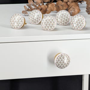Puede incluir: Un juego de seis pomos redondos para armarios con un patrón geométrico repetitivo de pequeños triángulos en tonos beige, blanco y cobre metálico. Cada pomo tiene una base de metal dorado y un acabado liso y brillante para muebles.