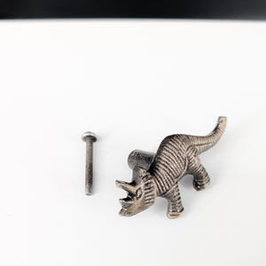 May include: A bronze-colored dinosaur-shaped cabinet knob with a matching screw. The knob is a detailed representation of a Triceratops, perfect for adding a unique touch to furniture or decor. The knob is made of metal.