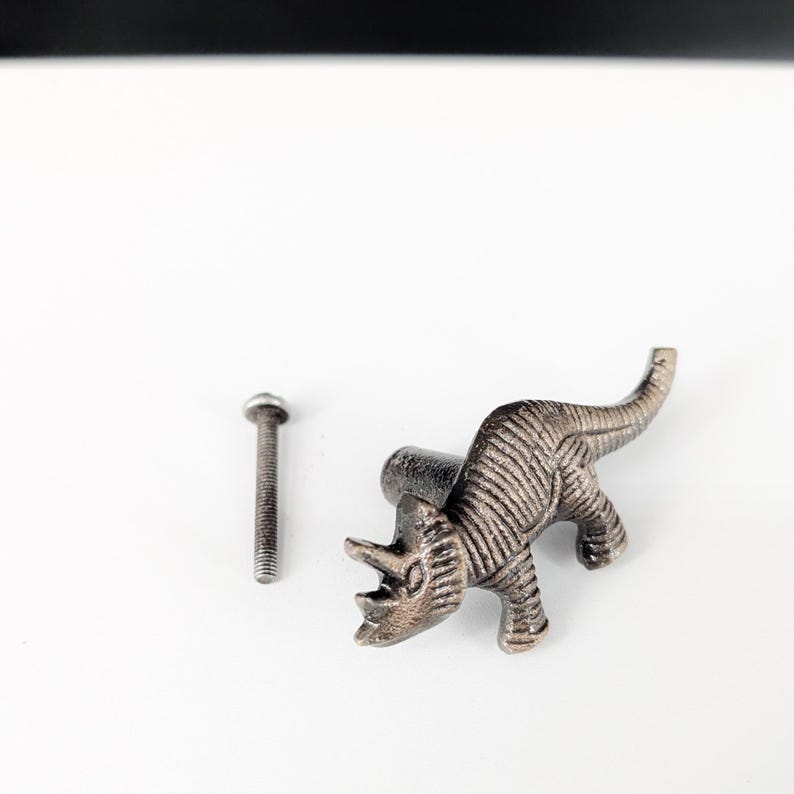 May include: A bronze-colored dinosaur-shaped cabinet knob with a matching screw. The knob is a detailed representation of a Triceratops, perfect for adding a unique touch to furniture or decor. The knob is made of metal.
