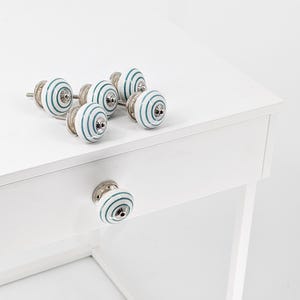 May include: Six white ceramic drawer knobs with teal blue stripes. The knobs have a silver finish and are arranged on a white surface.
