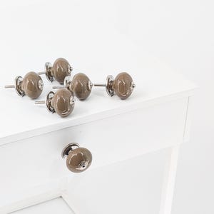 Peut inclure: Ensemble de six boutons de meubles en céramique marron avec des fixations en métal argenté. Les boutons ovales sont présentés sur une surface blanche, dont un est fixé à un tiroir blanc. Finition brillante.