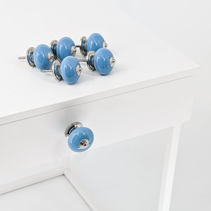 Può includere: Sei pomelli per armadi in ceramica blu chiaro con dettagli argentati. I pomelli sono disposti a semicerchio su una superficie bianca. Un pomello è montato su un cassetto bianco.