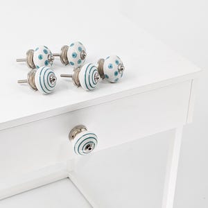 Könnte beinhalten: Satz mit sechs Keramik-Schubladengriffen mit weißer Basis und blau-weißen Mustern. Die Griffe haben eine silberne Metallbasis und eine Schraube. Die Griffe sind auf einer weißen Oberfläche abgebildet.