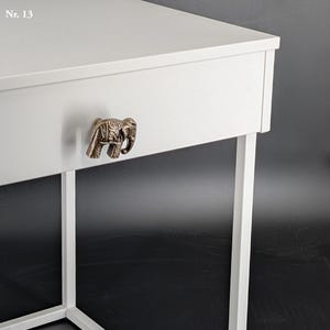 May include: A white side table with a drawer featuring a bronze elephant-shaped handle. The table has a minimalist design with clean lines and a sturdy, white metal frame. The text "Nr. 13" is in the upper left corner.