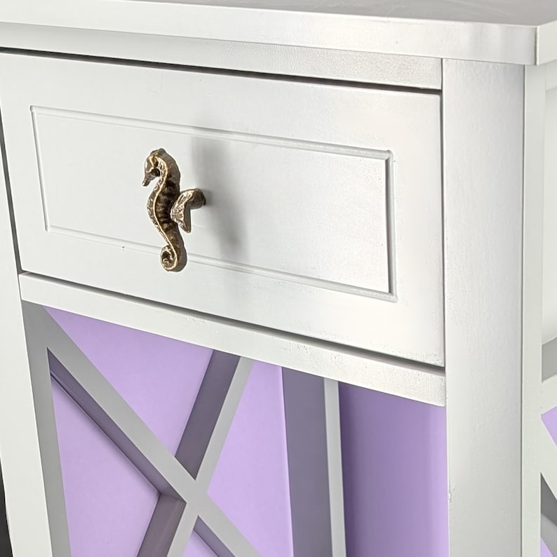 May include: White nightstand with a drawer featuring a seahorse-shaped handle. The drawer is set against a lavender background with a decorative X-shaped design. The nightstand has a coastal or nautical theme.