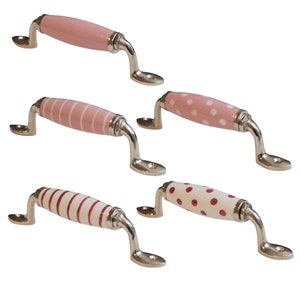 Puede incluir: Conjunto de cinco pomos de armario de cerámica con un diseño rosa y blanco o rojo y blanco. Los pomos tienen un acabado plateado y tienen forma de rectángulo redondeado.