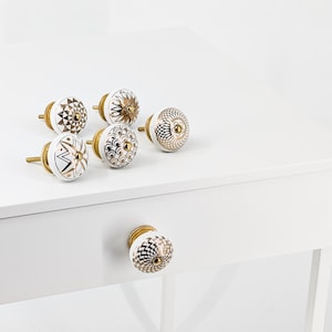 May include: Six round cabinet knobs featuring white ceramic bases with metallic gold and black geometric patterns. Each knob has a central gold-toned metal screw and a rounded top surface decorated with distinct intricate designs including stars, scales, and lines.