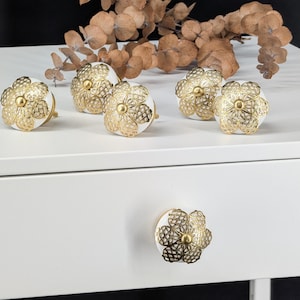 Könnte beinhalten: Ein Set aus sechs runden weißen Keramik-Möbelknöpfen, jeder mit einer goldfarbenen Metallauflage in Form einer fünfblättrigen Blume mit filigranen Details und einem kleinen zentralen Knopf, angeordnet auf einer weißen Oberfläche.