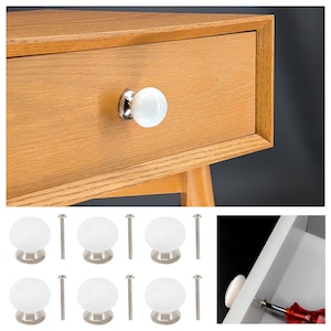 Könnte beinhalten: Set mit 8 weißen Keramik-Schrankknöpfen mit silberner Basis und Schrauben. Die Knöpfe sind rund und haben eine glatte Oberfläche.