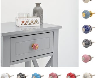 May include: Assortment of ceramic drawer knobs in various colors, including white, blue, pink, black, and red. The knobs have a floral design and a gold accent. The knobs are shown on a gray nightstand with a drawer.