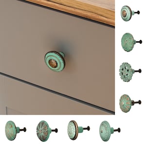 Könnte beinhalten: Sortiment von acht türkisfarbenen und bronzenen Schrankknöpfen mit verschiedenen verzierten Designs. Die Knöpfe sind auf einer grauen Schubladenfront und auf weißem Hintergrund abgebildet.