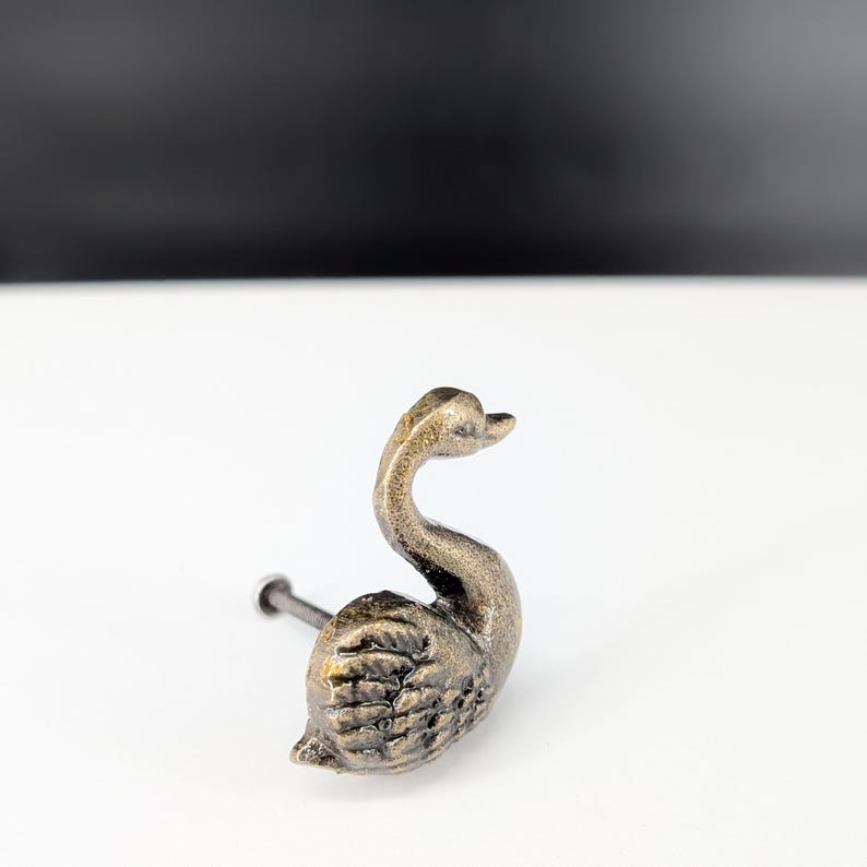 May include: A bronze-colored swan-shaped cabinet knob. The knob features a detailed swan design with a curved neck and textured feathers. The knob is mounted on a dark metal screw.