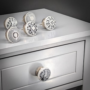 Op de afbeelding: Een verzameling decoratieve ladeknopen met diverse zwart-witte patronen. De knoppen zijn rond met een witte basis en zilverkleurige hardware. Ontwerpen omvatten bloemen, geometrische en abstracte patronen. Eén knop is bevestigd aan een grijze lade.