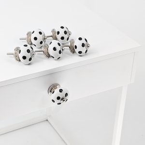 Op de afbeelding: Set van zes witte keramische kastknoppen met zwarte stippen. Elke knop heeft een zilveren metalen basis en een schroef.