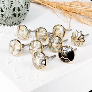 Op de afbeelding: Een verzameling van acht ronde kastknoppen met ingelegde parelmoersegmenten in goudkleurige metalen frames. Elke knop heeft een metalen bout met schroefdraad aan de achterkant en toont diverse geometrische patronen, waaronder ster-, honingraat- en gesegmenteerde cirkelvormige ontwerpen in wit-, crème- en zwarttinten.