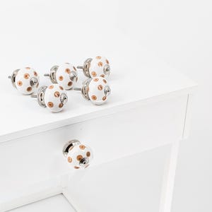 Peut inclure: Ensemble de six boutons de cabinet en céramique blanche avec des pois marrons. Les boutons ont une base en métal argenté et une vis.