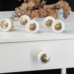 Könnte beinhalten: Ein Set aus sechs runden weißen Keramik-Möbelknöpfen, die jeweils mit einer dekorativen goldfarbenen Metalleinlage mit Blumenmuster auf der Vorderseite versehen sind. Ein Knopf ist an einer weißen Schublade befestigt, fünf liegen obenauf.