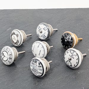 Könnte beinhalten: Satz mit 7 weißen Keramik-Schubladengriffen mit schwarzen und silbernen Akzenten. Die Griffe haben verschiedene Designs, darunter Uhren, Vintage-Text und ein Etikett für Olivenölseife.