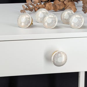 May include: A set of round cabinet knobs featuring a white center and a border of concentric gold dots. The knobs have a metallic gold base and are displayed on a white surface, with one knob attached to a drawer front and others arranged behind it.