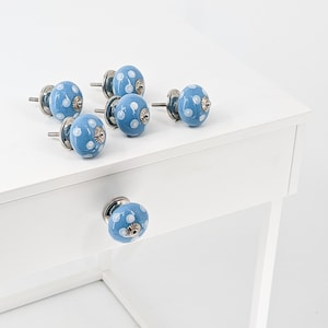 Op de afbeelding: Zeven blauwe keramische kastknoppen met witte stippen en zilveren accenten. De knoppen zijn gerangschikt op een wit oppervlak.