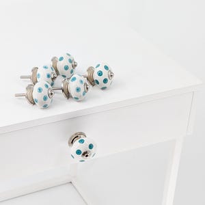 Peut inclure: Six boutons de tiroir en céramique blanche avec des pois turquoise et des ferrures argentées. Les boutons sont disposés sur une surface blanche. Un bouton est monté sur un tiroir blanc.
