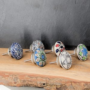 Op de afbeelding: Een verzameling keramische kastknoppen met diverse kleurrijke bloemen- en geometrische ontwerpen. De knoppen zijn wit met blauwe, groene, rode en zwarte patronen. Elke knop heeft een zilveren metalen schroef. Ze zijn gerangschikt op een houten oppervlak.