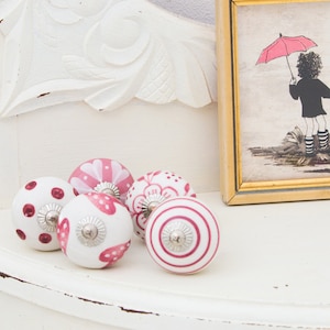 May include: Five pink and white ceramic drawer knobs with different floral and geometric patterns. The knobs are arranged on a white surface.