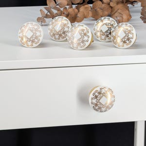 Può includere: Un set di sei pomelli rotondi per mobili con base in ceramica bianca e un motivo in filigrana color oro metallizzato. Cinque pomelli sono disposti su una superficie piana, mentre uno è fissato sulla parte anteriore di un cassetto bianco.