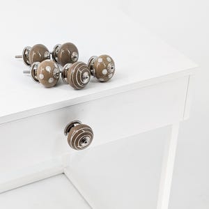 Könnte beinhalten: Satz von sechs braunen Keramik-Schrankknöpfen mit weißen Punkten und Wirbelmustern. Die Knöpfe haben eine silberne Metallbasis.