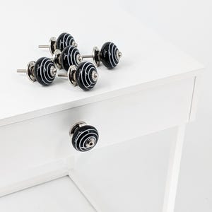 Könnte beinhalten: Sechs schwarze und weiße Keramik-Schubladengriffe mit silberner Beschläge. Die Griffe haben ein wirbelndes Muster und sind auf einer weißen Oberfläche angeordnet.