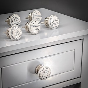 May include: A set of white ceramic cabinet knobs with silver-coloured hardware. Each knob has black text and star designs. The knobs are displayed on a light grey cabinet.