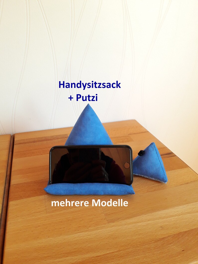 K&ouml;nnte beinhalten: Ein blauer Handyst&auml;nder aus Stoff in Form einer Pyramide. Der St&auml;nder steht auf einer Holzoberfl&auml;che. Der Text "Handysitzsack + Putzi" und "mehrere Modelle" ist im Bild zu sehen.