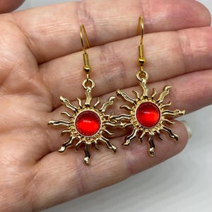 May include: A pair of gold-colored sunburst earrings with red centers. The earrings have a hook closure and are displayed against a light background. The sunburst design features radiating rays around a circular red center.