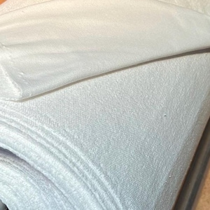 Pode incluir: Um rolo de tecido branco, provavelmente uma toalha ou material para roupão, está enrolado e preso com uma faixa branca. O tecido tem uma superfície macia e texturizada.