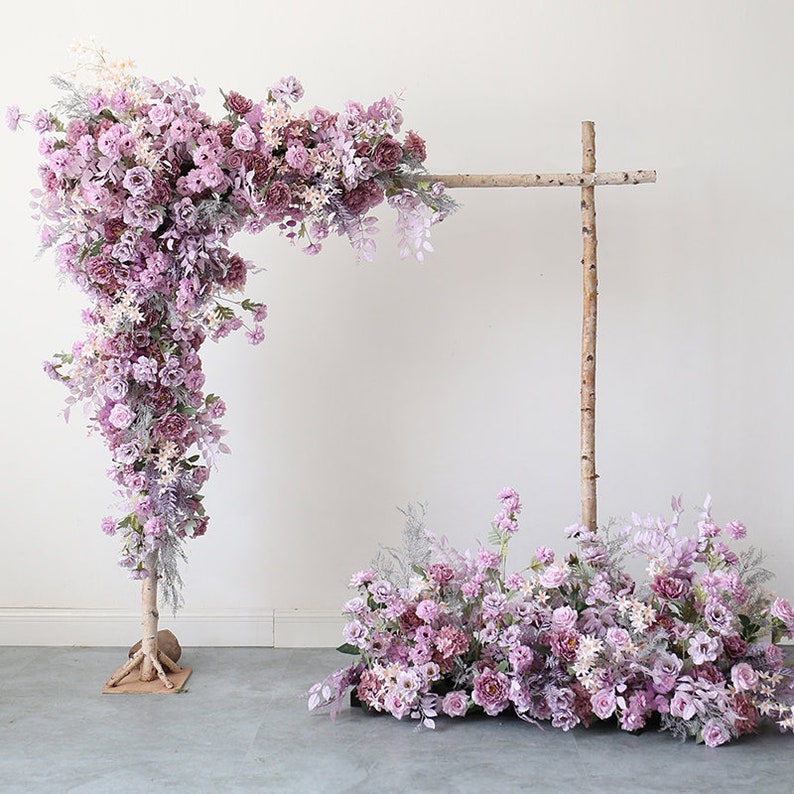 May include: Two floral arrangements with purple flowers and greenery. One arrangement is a cascading floral arch with a birch wood frame. The other arrangement is a smaller, more compact arrangement on the ground.