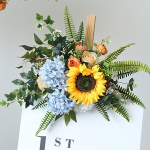 May include: A floral arrangement with a sunflower, blue hydrangeas, and orange roses. The arrangement is attached to a wooden sign with the text "1ST" on it.