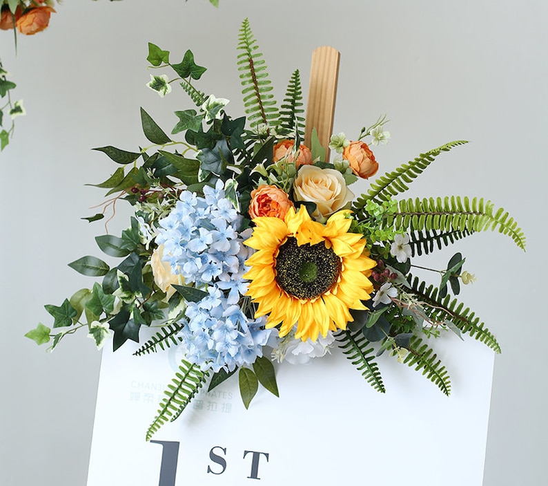 May include: A floral arrangement with a sunflower, blue hydrangeas, and orange roses. The arrangement is attached to a wooden sign with the text "1ST" on it.