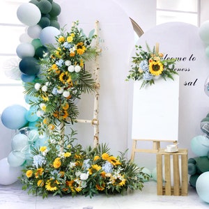 May include: A white easel with a blank canvas, a wooden crate, and a floral arrangement with sunflowers, hydrangeas, and greenery. The floral arrangement is on a white ladder and on the floor. There are blue and green balloons in the background.