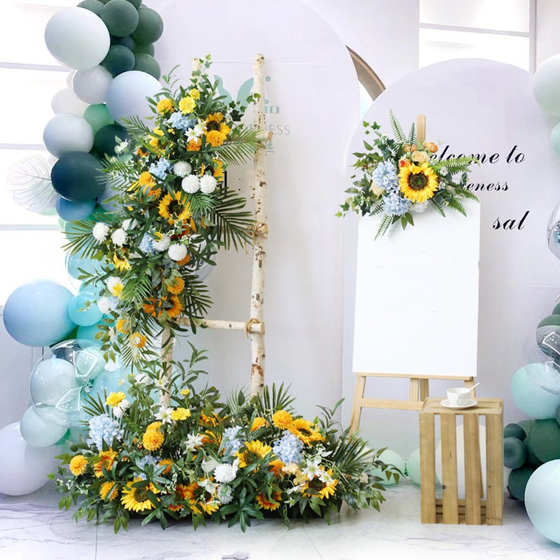 May include: A white easel with a blank canvas, a wooden crate, and a floral arrangement with sunflowers, hydrangeas, and greenery. The floral arrangement is on a white ladder and on the floor. There are blue and green balloons in the background.