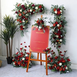 May include: A red welcome sign with gold trim on an easel surrounded by artificial flower arrangements with red, white, and orange flowers and green leaves.