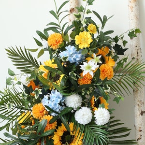 May include: A floral arrangement featuring sunflowers, white and blue hydrangeas, and other yellow flowers. The arrangement is set against a white background and includes green leaves.