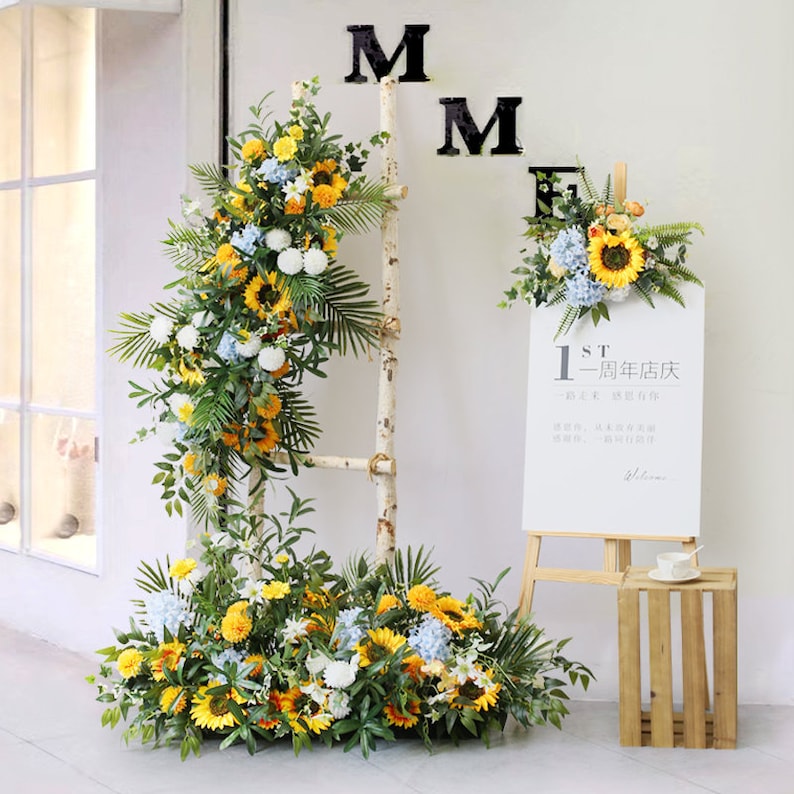 May include: A white wooden ladder decorated with a floral garland of yellow sunflowers, white pom poms, blue hydrangeas, and green leaves. The garland is also draped on the floor in front of the ladder. A white easel with a sign that reads "1st - 周年店庆" is to the right of the ladder. A small wooden crate is in the bottom right corner.