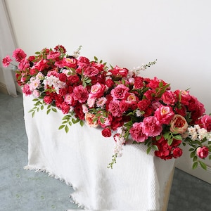 May include: A long garland of pink and red artificial flowers, including roses and carnations, arranged on a white tablecloth. The garland is perfect for a wedding or other special event.