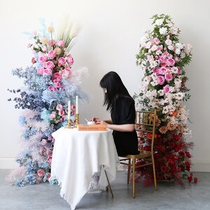May include: A white table with a white tablecloth and a book titled "Gasta Laetitia" on top. A person is sitting at the table, and there are two tall floral arrangements behind them. One arrangement is mostly pink and white flowers, and the other is mostly blue and pink flowers.